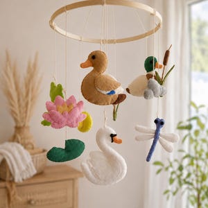 May include: A hanging mobile featuring felt figures suspended from a circular wooden frame. The figures include a brown duck, a mallard duck with cattails, a white swan, a pink water lily with a green leaf, and a blue and white dragonfly.