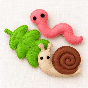 May include: A set of three felt craft pieces including a bright green oak leaf, a pink worm with two black circular eyes, and a beige snail with a dark brown spiral shell and two black eyes, all featuring visible stitched edges.
