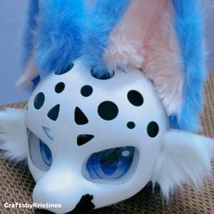 Peut inclure: T&ecirc;te d'animal en fourrure blanche avec des oreilles en fourrure bleue et rose. La t&ecirc;te a des taches noires et un motif triangulaire noir sur le visage. Les yeux sont bleus et ont un rev&ecirc;tement en plastique transparent.