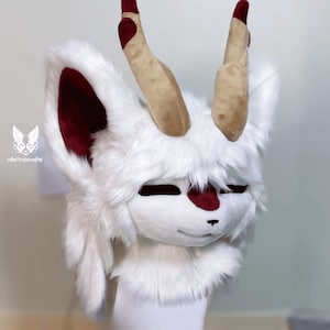 May include: White furry animal head with red ears and two long, tan horns. The head has a closed mouth and a small, red nose.