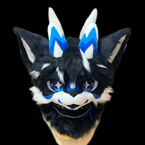 May include: A black and white furry fursuit head with blue accents. The head features large, expressive eyes, blue markings, and white horns with blue details. A silver chain is around the mouth area.