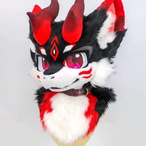 May include: A detailed furry head with red horns, ears, and accents. The face is primarily white with black and red markings, and large pink eyes. A brown collar is around the neck. The head is mounted on a mannequin head.