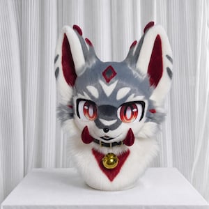 May include: A plush animal head mask featuring grey, white, and deep red faux fur. The mask has large, expressive red eyes, pointed ears with red accents, two red fangs, and a black collar with a large, shiny gold bell hanging at the center.