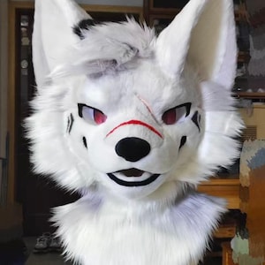 May include: A white furry fox head with large, upright ears and red eyes. The fox head has black markings around the eyes and a black nose. A red line is painted across the nose. The head is part of a costume.