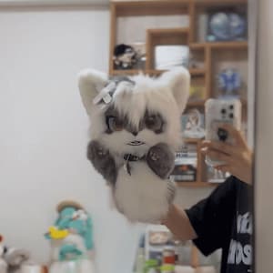 May include: A plush, furry animal head with large eyes, white fur, and gray accents. The toy has a small bow and a collar. The plush is being held up in front of a mirror, with a person's arm and a phone visible.