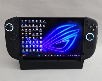 May include: Black handheld gaming device with a screen displaying a blue and purple logo. The device has buttons and joysticks on either side. It is docked in a black stand.