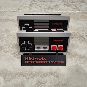 Op de afbeelding: Twee grijze en zwarte Nintendo Entertainment System-controllers met rode knoppen worden weergegeven op een zwarte standaard met de tekst "Nintendo Entertainment System".
