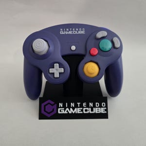 May include: A purple Nintendo GameCube controller with a white analog stick, a yellow C-stick, and colored buttons. The controller is on a black stand with the Nintendo GameCube logo and text.