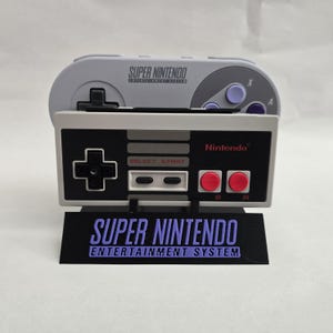 Pode incluir: Uma réplica do controle e console do Super Nintendo Entertainment System. O controle cinza fica em cima de uma réplica de console em preto e branco com botões vermelhos. A base diz "SUPER NINTENDO ENTERTAINMENT SYSTEM" em roxo.