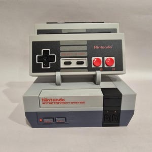 May include: A gray and black Nintendo Entertainment System (NES) replica. The console features a controller design on top with red buttons and the Nintendo logo. The bottom section has the Nintendo Entertainment System logo and cartridge slots.