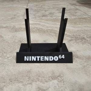 Puede incluir: Soporte de plástico negro para cartuchos de videojuegos con el texto "NINTENDO 64" impreso en la parte delantera.