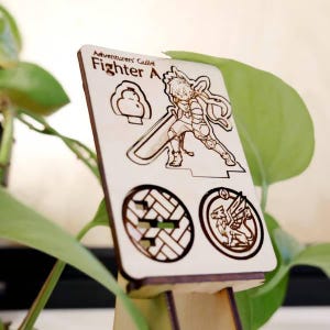 May include: Wooden laser-cut card featuring a warrior illustration and the text "Adventurers' Guild Fighter A". The card includes additional etched designs of a cloud, a geometric pattern, and a griffin. The card is displayed in a wooden stand.