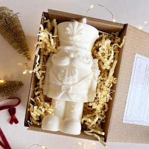 Könnte beinhalten: Eine detaillierte weiße Kerze in Form eines Nussknacker-Soldaten, präsentiert in einer braunen Schachtel mit goldenem Papier. Die Kerze hat einen Schnurrbart, eine Mütze und Uniformdetails. Lichterketten und kleine Weihnachtsbäume sind im Hintergrund.