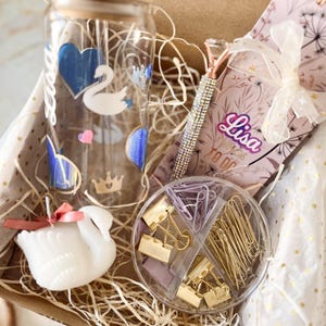 Puede incluir: Una caja de regalo que contiene un vaso de vidrio con diseños de cisnes y corazones, una vela con forma de cisne, un cuaderno, un bolígrafo y clips. El vaso tiene el nombre "Lisa". La caja está llena de paja decorativa.
