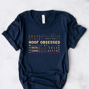 May include: A navy blue t-shirt with a graphic design featuring the words "HOOF OBSESSED" in gold lettering and other related words in pink and orange lettering.