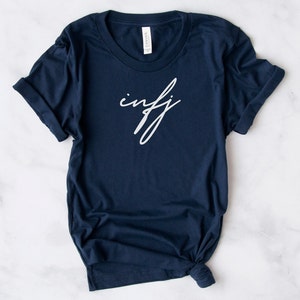May include: Navy blue t-shirt with the white cursive text "infj". The shirt is made of a soft material and has a classic crew neck. The sleeves are rolled up, and the bottom is knotted.
