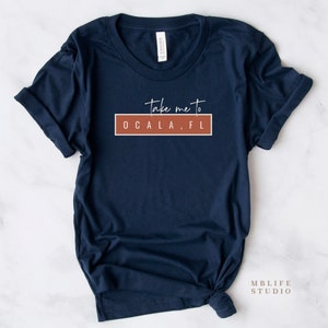 May include: Navy blue t-shirt with the text "take me to OCALA, FL" in white script and block letters, set against a rust-colored rectangle. The shirt is folded and knotted at the bottom.
