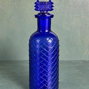 May include: A cobalt blue glass bottle with a diamond-patterned textured surface and a matching stopper. The stopper features a rectangular top with a spiked edge and the word POISON embossed in the centre. The bottle has a cylindrical body.
