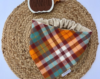 May include: A tartan dog bandana in orange, teal, and burgundy, with a beige elasticated band. A decorative pumpkin with the word "THANKFUL" is above the bandana, along with a fall leaf. The bandana is on a woven mat.