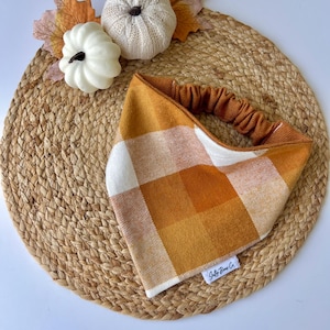 May include: A fall-themed headband with a plaid pattern in shades of orange, white, and brown. The headband is set on a woven mat with decorative pumpkins and leaves.