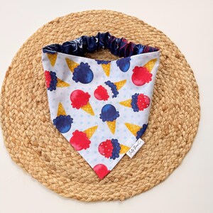 May include: A white dog bandana with a repeating pattern of red and blue ice cream cones on a light blue polka dot background. The bandana has a navy blue trim with a star pattern. The tag reads "Salty Bean Co."