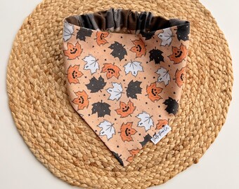 May include: A Halloween-themed dog bandana featuring a peach background with orange jack-o'-lantern leaves, black leaves, and white ghost leaves. The bandana has a black and white plaid pattern on the reverse side.