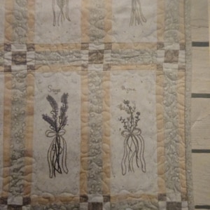 May include: A quilted fabric runner featuring a grid pattern with alternating blocks. The blocks display embroidered herb bundles tied with ribbons, labeled with the text Sage and Thyme. The color palette includes cream, yellow, and brown tones.