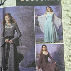 May include: A Simplicity Costumes sewing pattern envelope featuring three floor-length dresses with long bell sleeves. The pattern displays a dark grey dress, a light blue dress with sheer sleeves, and a dark brown dress, all with medieval-inspired designs.