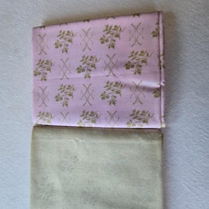 Pode incluir: Duas peças de tecido dobradas. A peça superior é rosa com um padrão floral dourado e um design repetitivo. A peça inferior é bege claro com um padrão floral subtil. Os tecidos são provavelmente para costura ou artesanato.