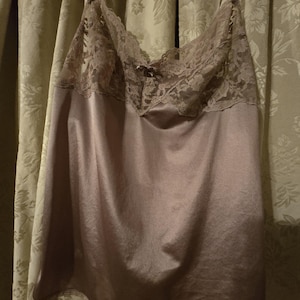 May include: A mauve-colored camisole with delicate lace trim at the neckline and straps. The camisole is made of a silky material and hangs from a metal hanger. The lace features a floral design.