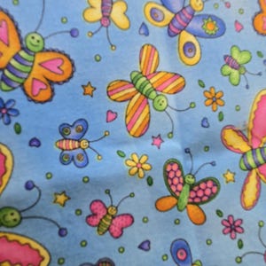 May include: A light blue fabric with a repeating pattern of colorful butterflies, flowers, stars, and hearts. The butterflies have orange, pink, yellow, and blue wings with green striped bodies. The design is whimsical and cheerful.
