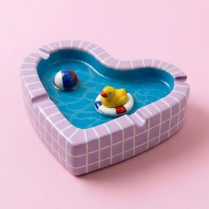 Peut inclure: Cendrier en forme de cœur avec un motif de piscine. Le cendrier présente un intérieur bleu avec un petit ballon de plage et un canard en caoutchouc dans une bouée de sauvetage. L'extérieur est lavande avec un motif de grille blanche. Le fond est rose.
