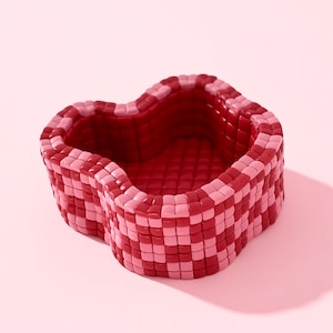 Puede incluir: Un cuenco decorativo en forma de corazón en tonos rojos y rosas. El cuenco tiene un aspecto tejido con un patrón de cuadros. El interior es de un rojo más oscuro sólido, mientras que el exterior presenta cuadrados alternos de rosa claro y rojo.