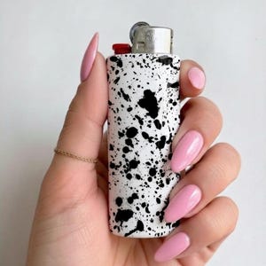 Puede incluir: Un encendedor blanco con diseño de pintura de salpicaduras negras. El encendedor se sostiene en una mano con esmalte de uñas rosa y un anillo dorado. El encendedor tiene una parte superior plateada y un botón rojo.