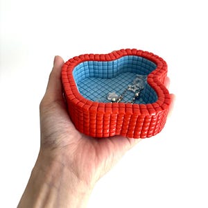 May include: A red and blue decorative jewelry dish shaped like a swimming pool. The dish has a red, textured exterior and a blue interior with a tiled pattern. A silver necklace with a star charm rests inside the dish.
