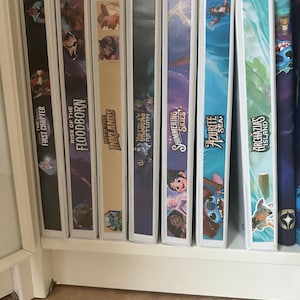 Puede incluir: Una colección de carpetas blancas con ilustraciones coloridas y texto. Los títulos incluyen "The First Chapter", "Rise of the Floodborn" e "Into the Inklands". Las carpetas están en un estante blanco.