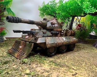 May include: A detailed model tank in shades of brown, green, and tan, with a large cannon and a front blade. The tank sits on a bed of green moss, with artificial plants in the background.