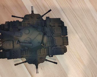 May include: A dark gray and brown toy tank with a detailed design. The tank has a long barrel and a turret with a small hatch. The tank is sitting on a wooden surface.