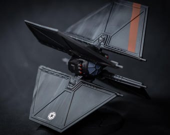 May include: A black TIE Interceptor model featuring angular solar array wings with a single vertical orange stripe on the upper wing. The central cockpit sphere has a circular window, and the lower wing displays a small white Imperial emblem.