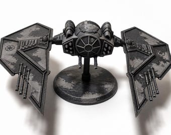 May include: A detailed, dark gray model of a TIE fighter spacecraft, featuring distinctive wing panels and a central cockpit. The model is mounted on a circular base, showcasing intricate design elements and a weathered finish.