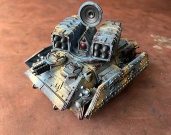May include: A miniature armored tank model painted in light blue with heavy weathering and rust effects. The vehicle features two missile launchers, a central radar dish, a small turret with a gatling gun, and the word ARMAGEDDON on the side panel.