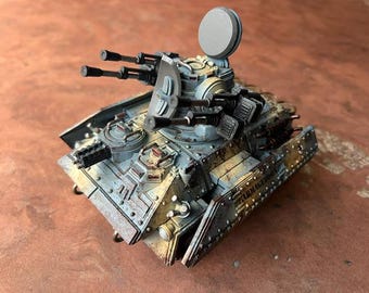 May include: A miniature scale model of a military tank painted in light blue and weathered tan tones. The tank features a rotating turret with dual mounted cannons, a circular hatch, and side armor plates with the word ARMAGEDDON embossed on them.