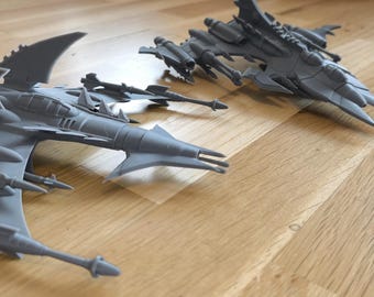 May include: Two gray, futuristic spaceship models with intricate details, including wings, spikes, and multiple weapon mounts. The models are displayed on a wooden surface, showcasing their design and structure.