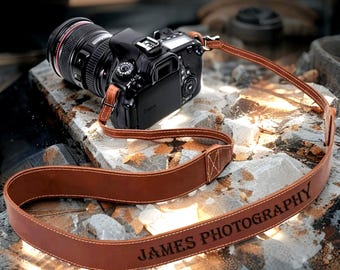 Może przedstawiać: Brązowy skórzany pasek do aparatu z napisem "JAMES PHOTOGRAPHY" jest przymocowany do czarnego aparatu DSLR. Pasek jest spersonalizowany i przeszyty białą nicią. Tekst "Personalized Leather Camera Strap" znajduje się na górze obrazu.