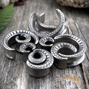 May include: An assortment of silver-toned ear gauges in various sizes and spiral designs. The gauges feature intricate patterns and are displayed on a weathered wooden surface, showcasing a rustic aesthetic. The jewelry is designed for stretched ear piercings.