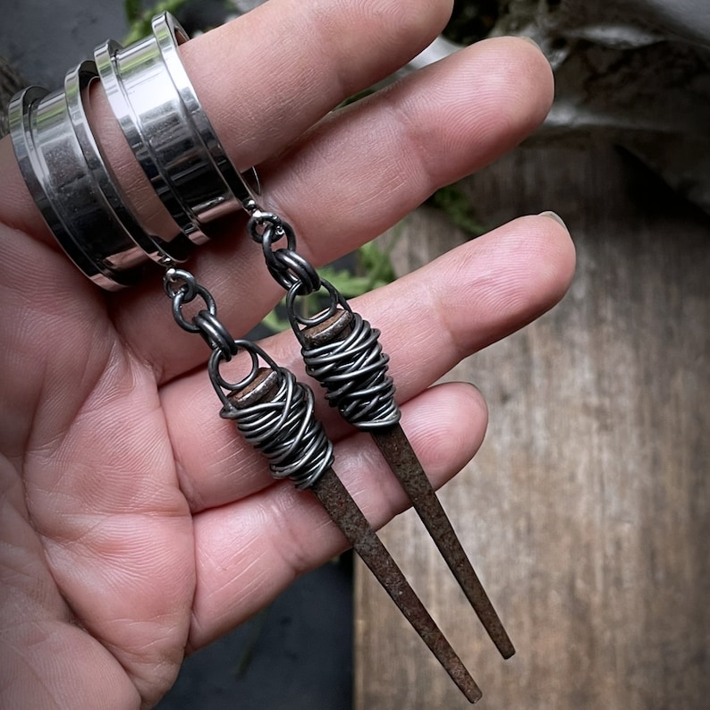 May include: A pair of silver earrings with long, thin, wire-wrapped, nail-shaped pendants. The nails are wrapped in silver wire and have a dark, aged finish.