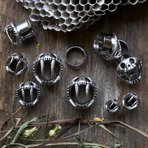May include: A set of silver metal ear gauges with a variety of designs, including a skull with fangs, a pair of fangs, and a simple circular design. The gauges are displayed on a wooden surface with a honeycomb pattern.
