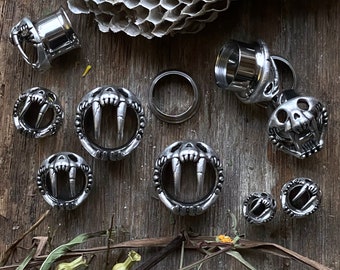 Può includere: Un set di dilatatori per orecchie in metallo argentato con una varietà di design, tra cui un teschio con zanne, una coppia di zanne e un semplice design circolare. I dilatatori sono esposti su una superficie di legno con un motivo a nido d'ape.
