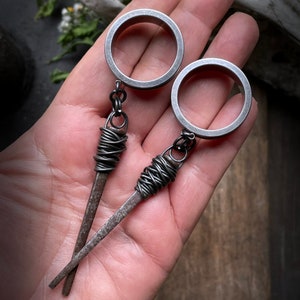 May include: Two silver metal ear gauges with a wire wrapped around the end of each gauge, resembling a nail. The gauges are hanging from a hand.