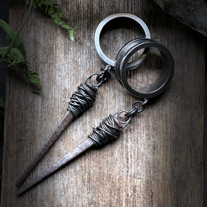 May include: A silver key chain with two metal rings and two wire-wrapped nails. The nails are attached to the rings with a silver chain.